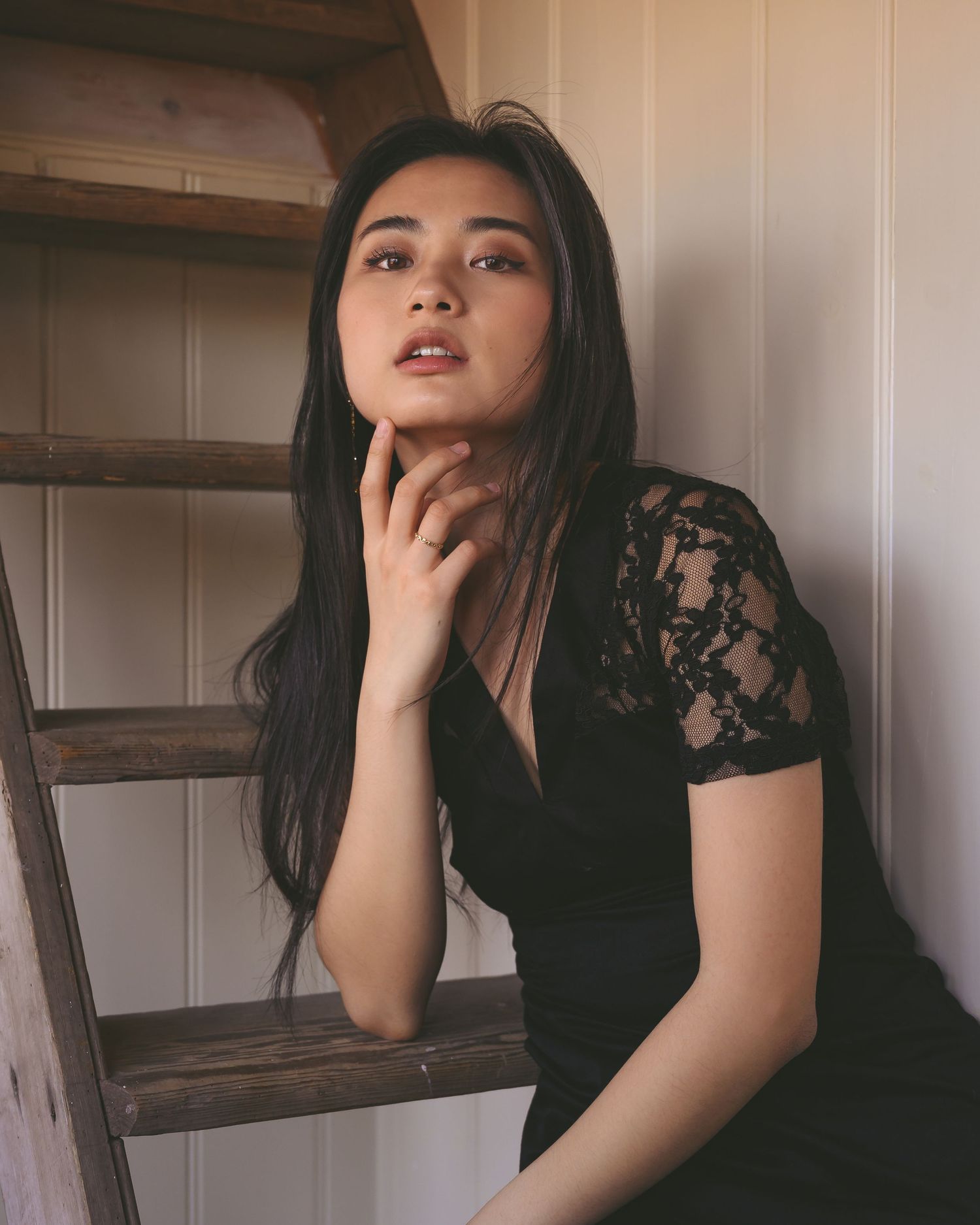 Young woman in black lace dress sits thoughtfully by a wooden ladder in a softly lit interior.