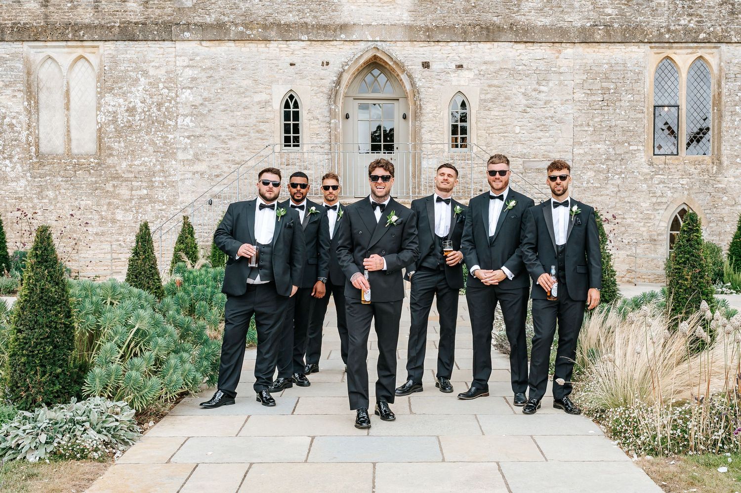Groom and groomsmen in matching black tuxedos pose confidently outside a historic stone church.