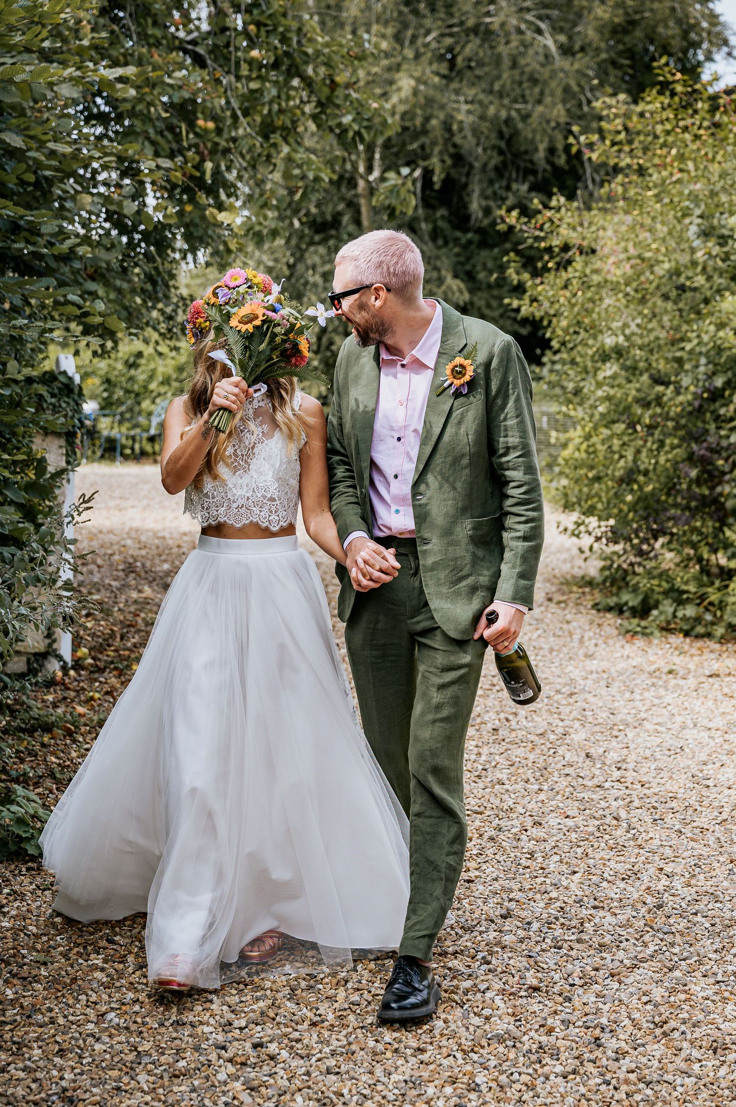 Bride and groom walk hand in hand on gravel path, bride holding vibrant wildflower bouquet.