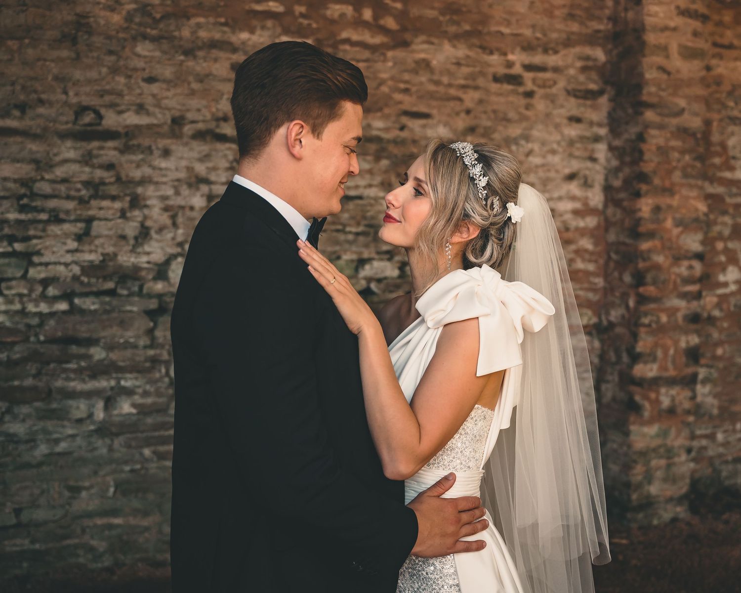 Bride and groom share a romantic moment in front of a rustic brick wall, bride wearing a white gown and veil.