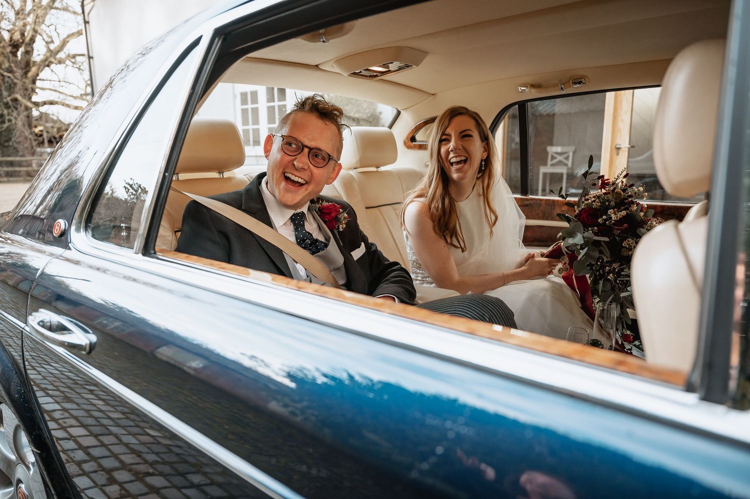Happy bride and groom laugh together inside a classic vintage car on their wedding day.