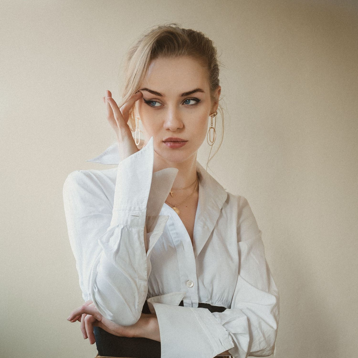 Elegant blonde woman in white blouse posing with hand near face against neutral background.