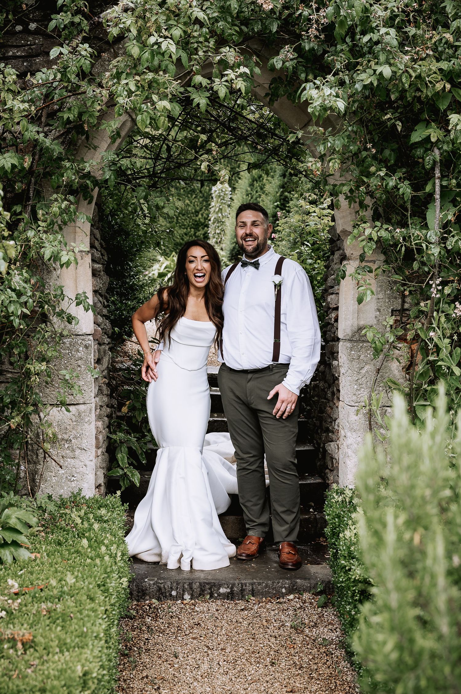 Joyful bride and groom posing under a lush green garden archway on their wedding day.