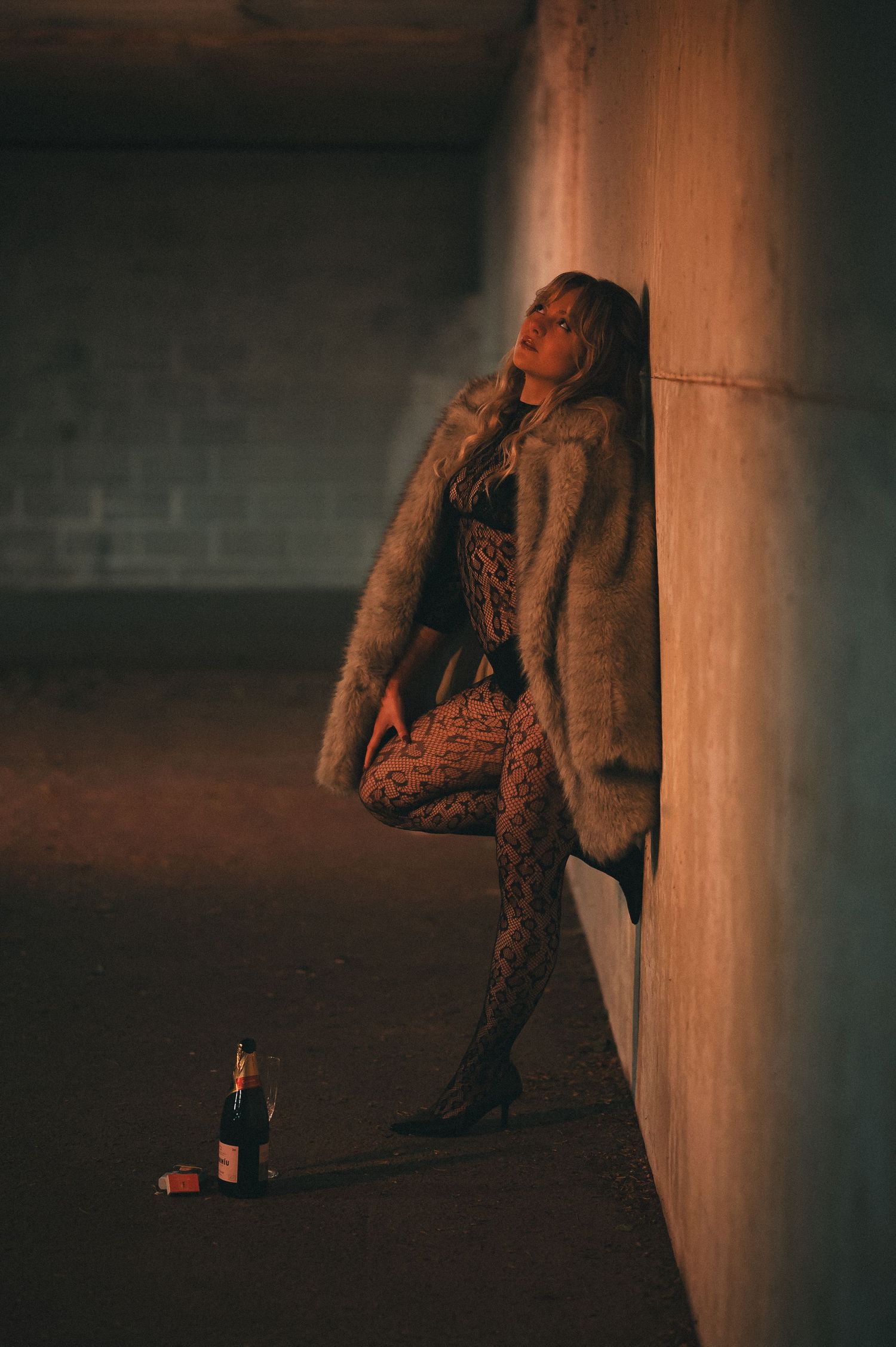 Woman in fur coat and leopard print leggings leaning against concrete wall with wine bottle.