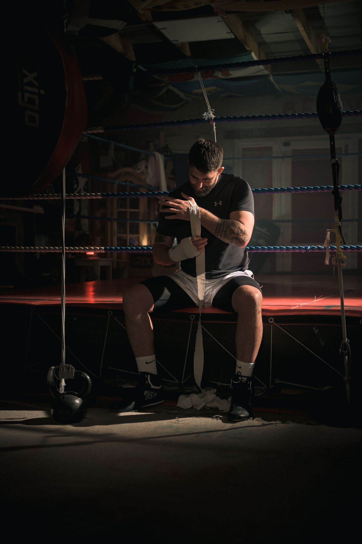 Male boxer wrapping hands while sitting in corner of dark boxing ring gym.