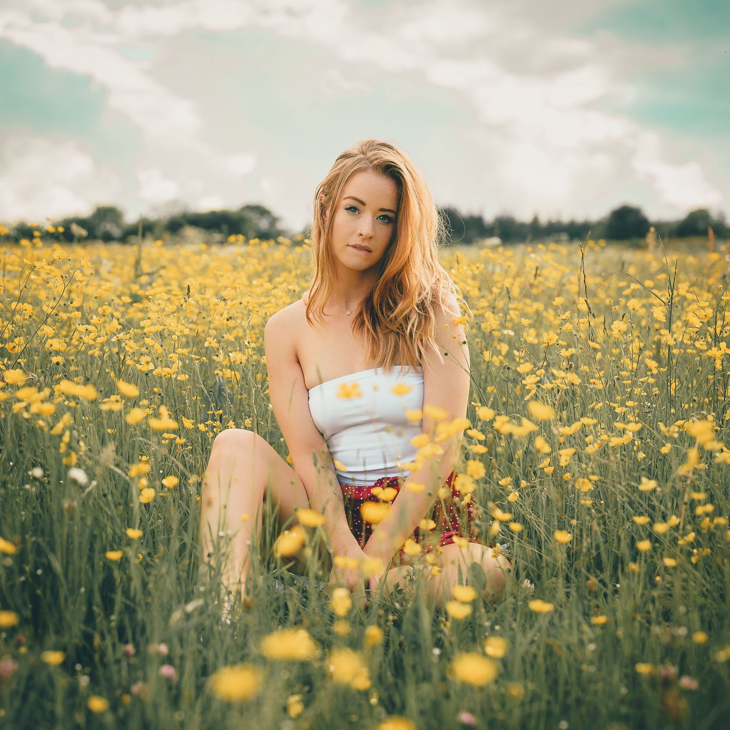 Young blonde woman sitting in a vibrant yellow wildflower field wearing a white top.