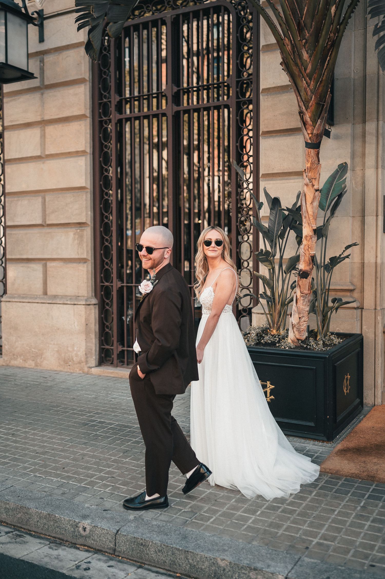 Stylish bride and groom in sunglasses pose outside a grand building with palm trees in Barcelona.