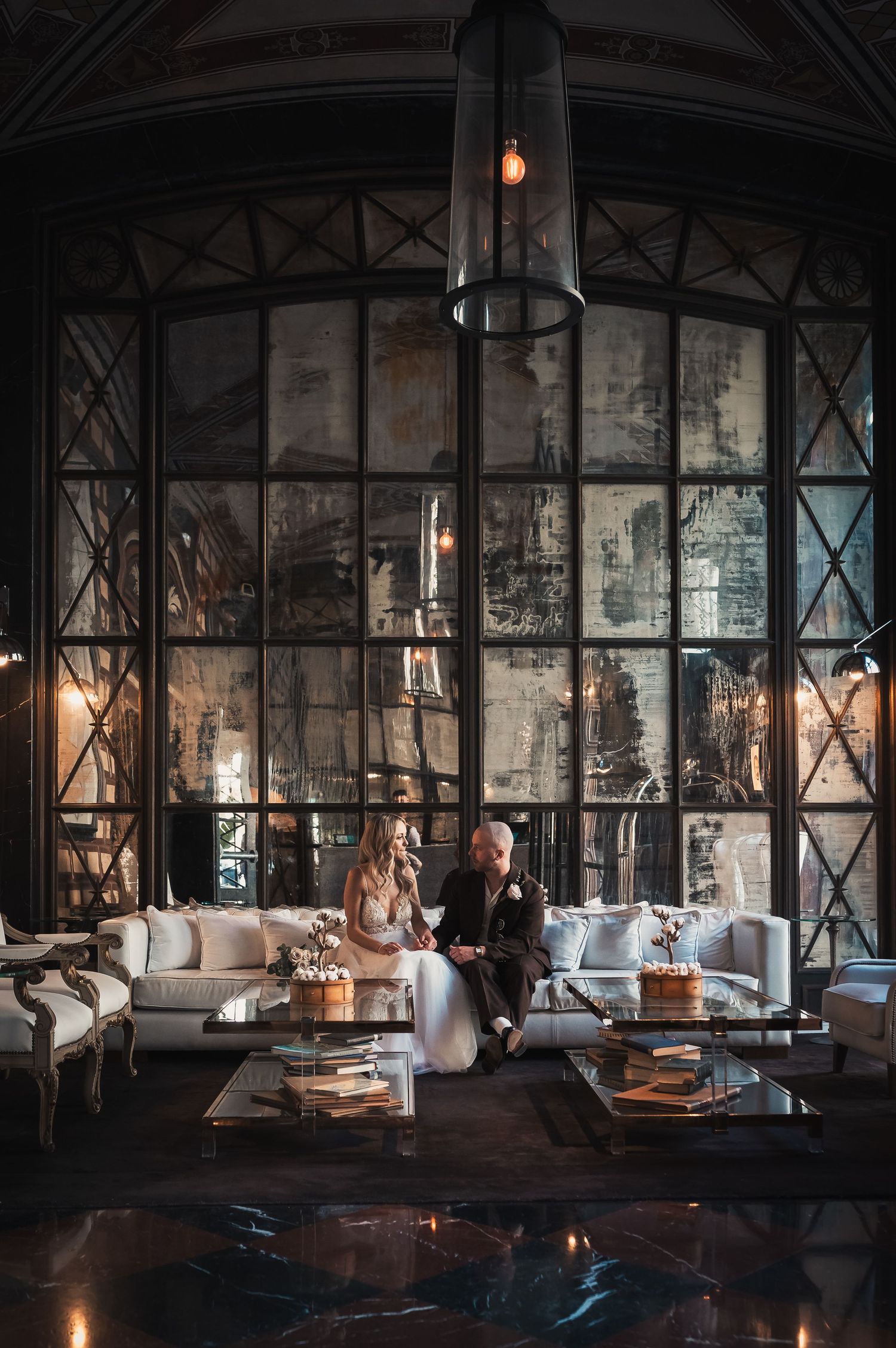 Bride and groom seated together in luxurious dark interior with tall arched windows.