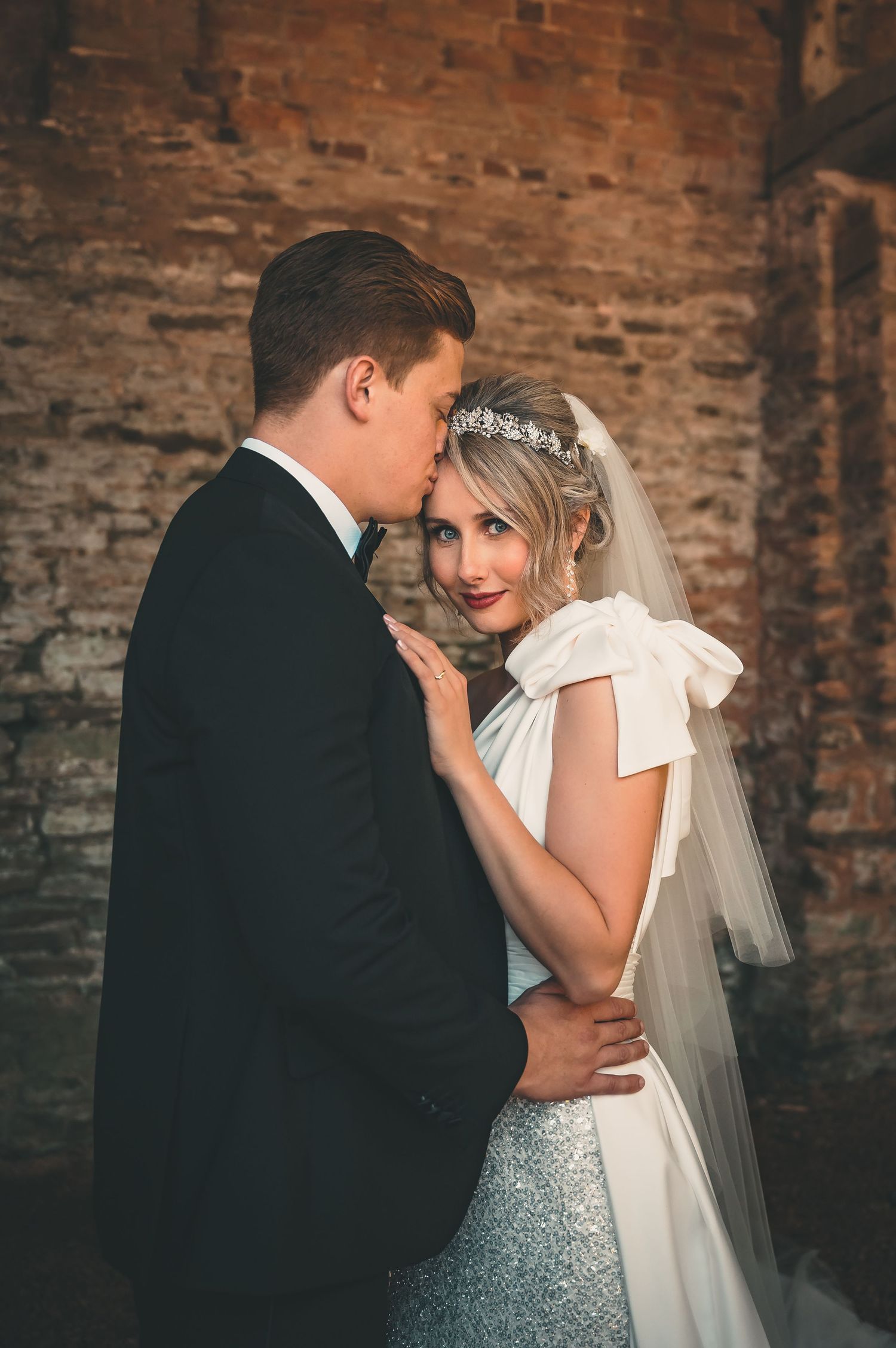 Groom kissing bride wearing crystal tiara and bow veil against rustic brick wall.