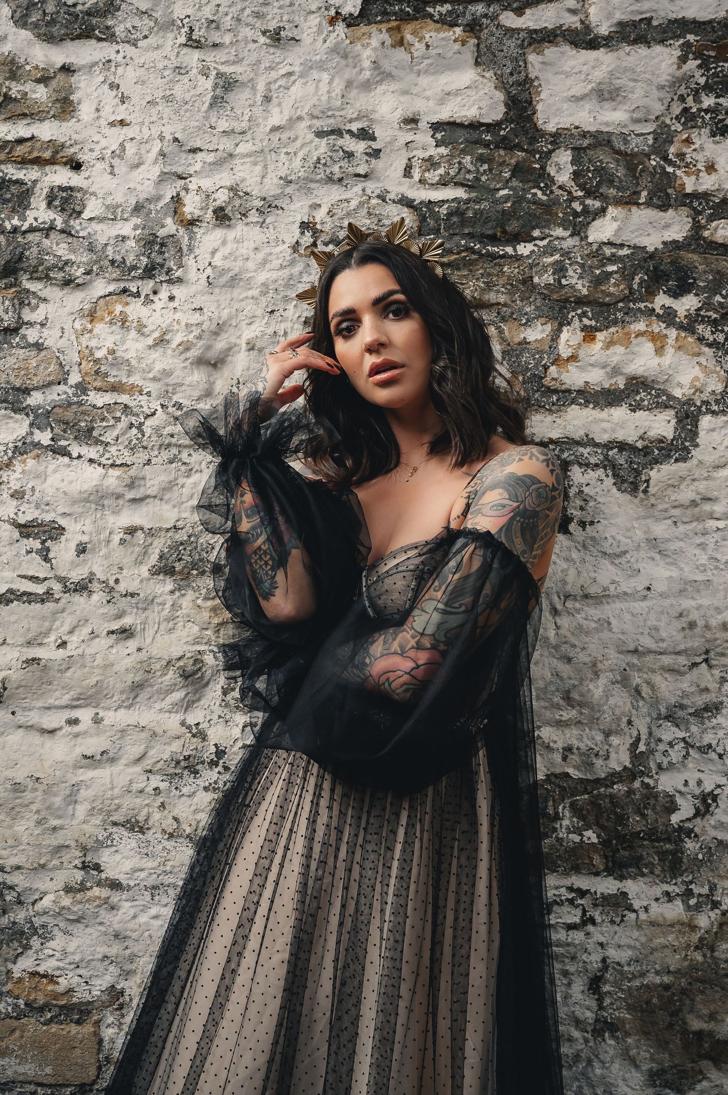 Tattooed woman in black tulle gown with gold crown posing against rustic stone wall.
