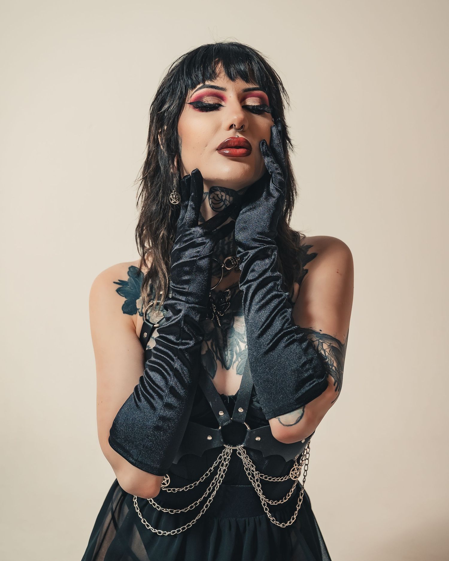 Gothic woman in black gloves and chains with bold red makeup posing against beige background.
