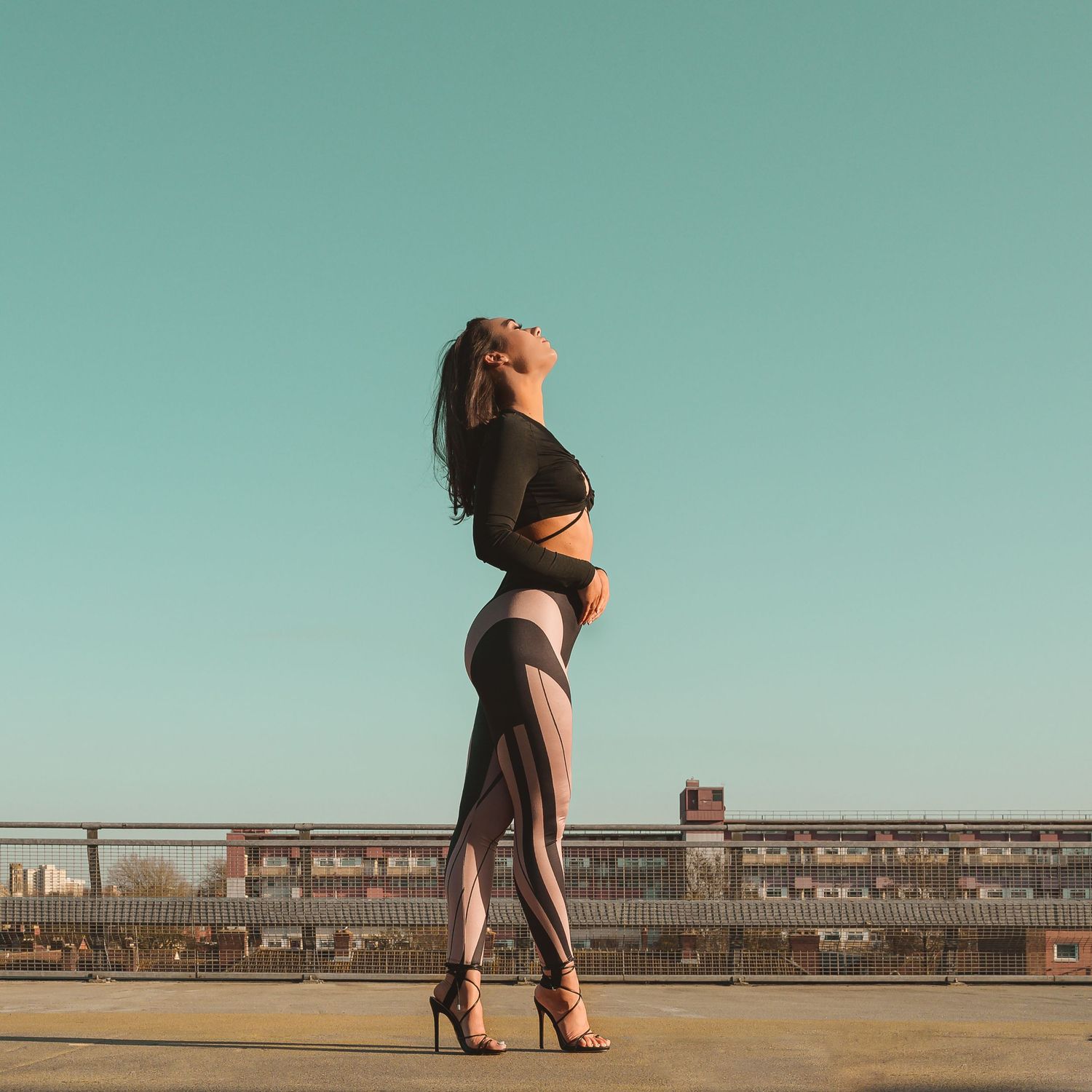 Woman in black crop top and sheer pants posing on rooftop against clear blue sky.