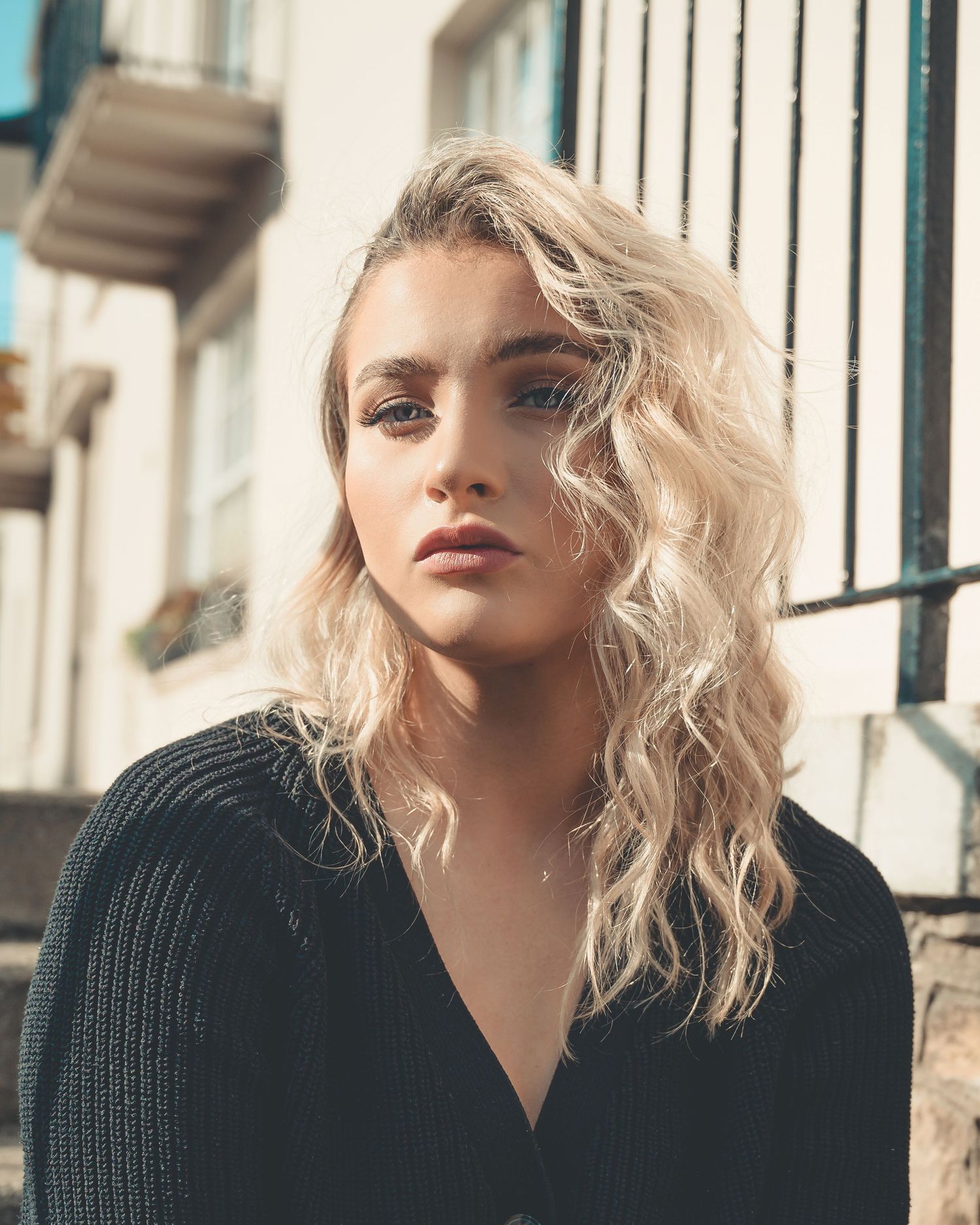 Blonde woman with wavy hair in black cardigan poses outdoors in warm sunlight.