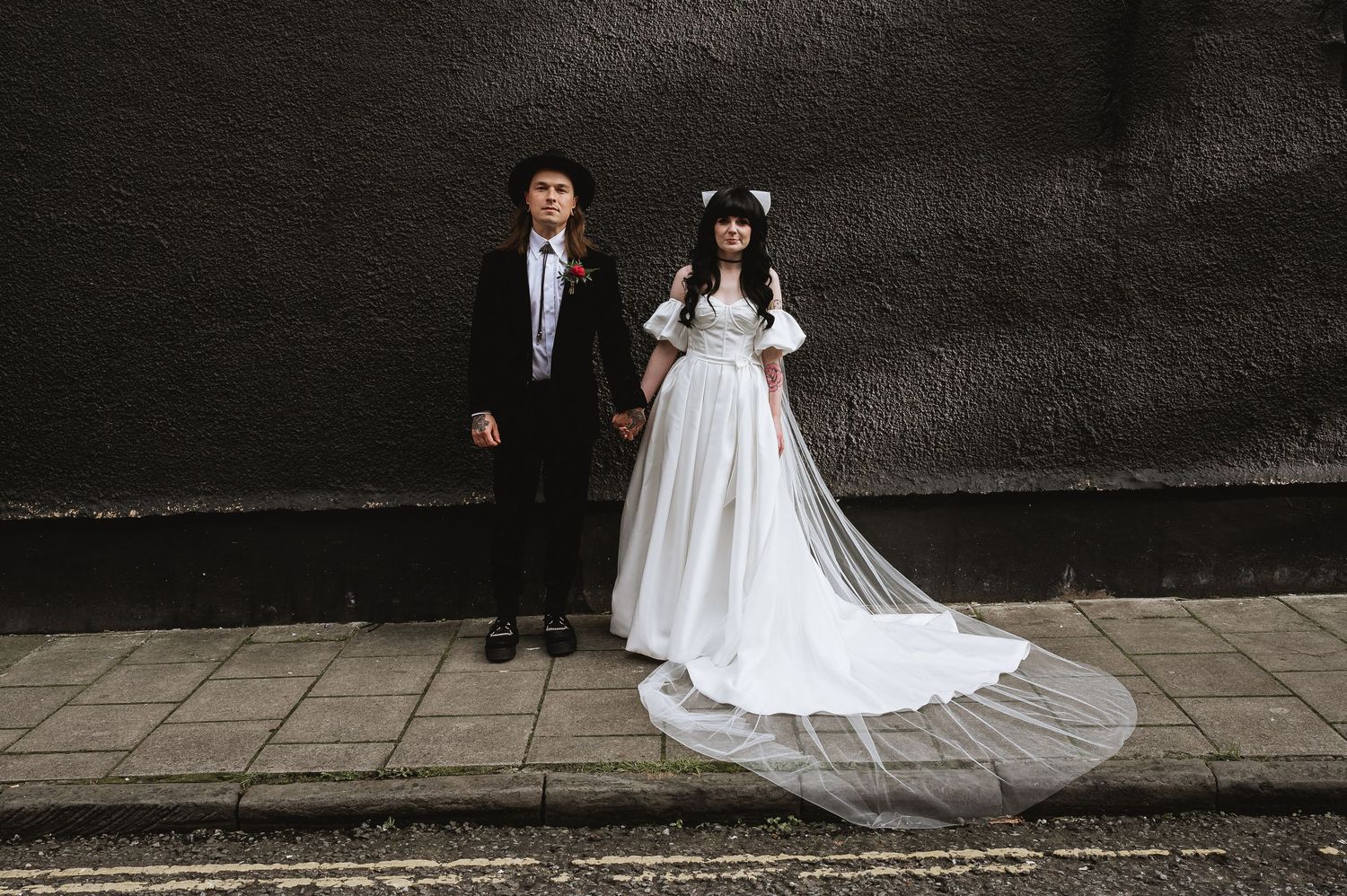 Wedding couple holding hands in front of dark wall, bride in flowing white dress with cathedral veil.