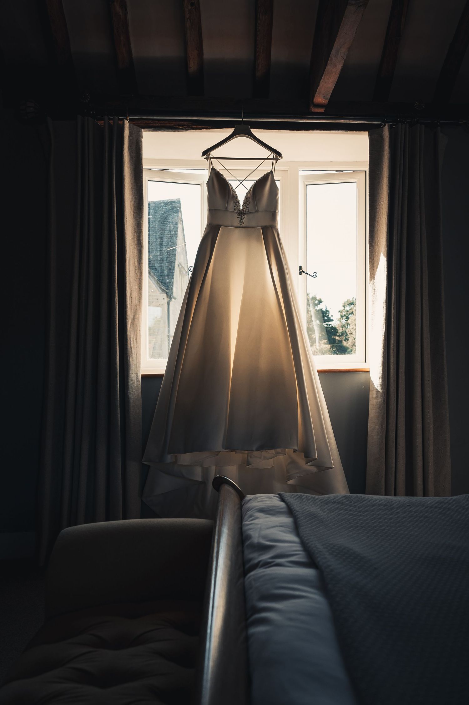 Elegant A-line wedding dress hanging in a window, backlit by soft natural light in a bedroom.