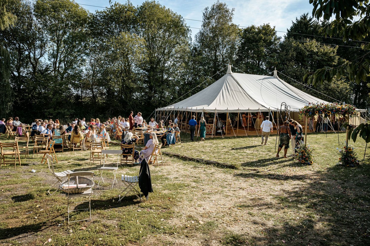 Guests gather outside a large white sailcloth wedding tent on a sunny green outdoor venue.