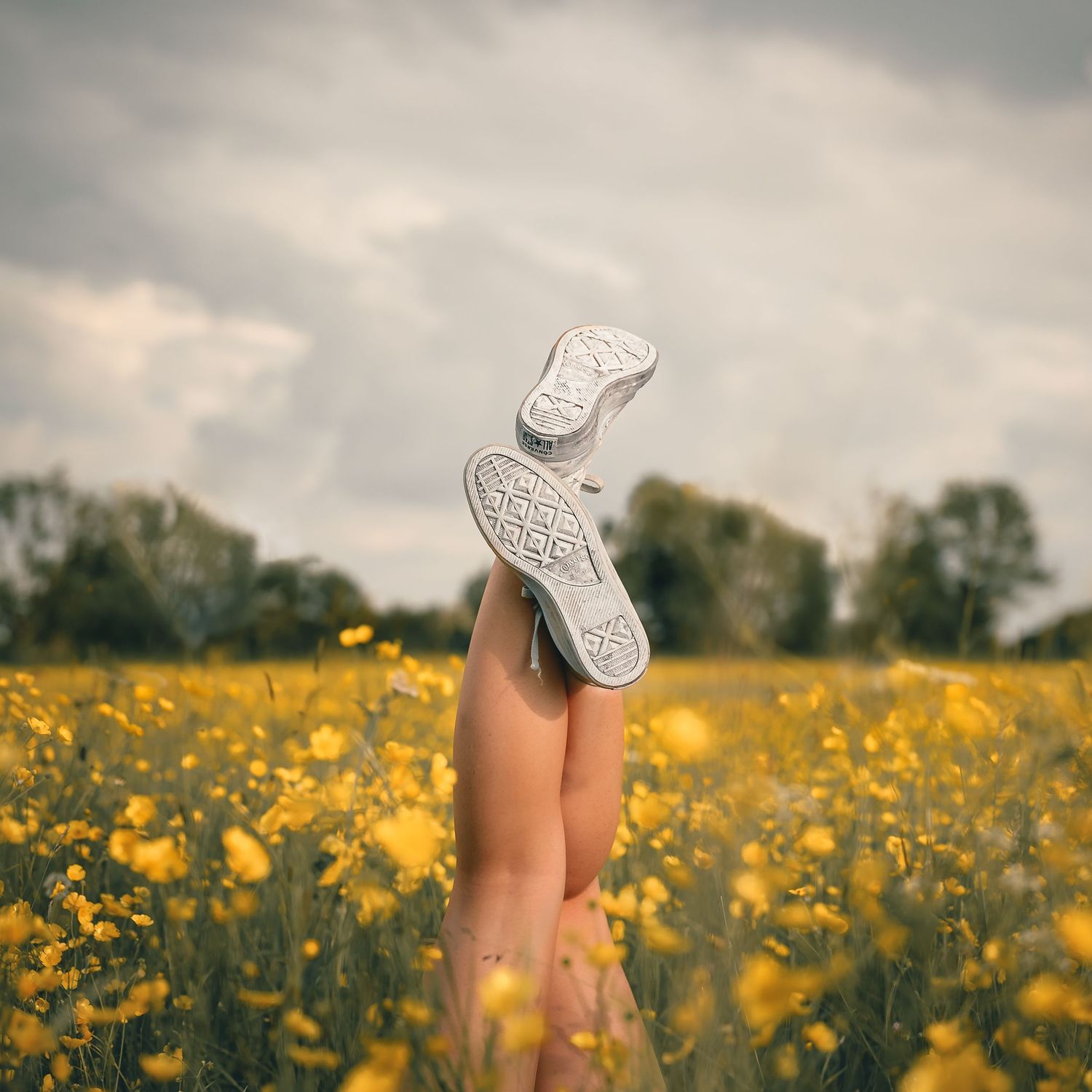 Legs raised wearing white sneakers above a field of yellow flowers under cloudy sky.