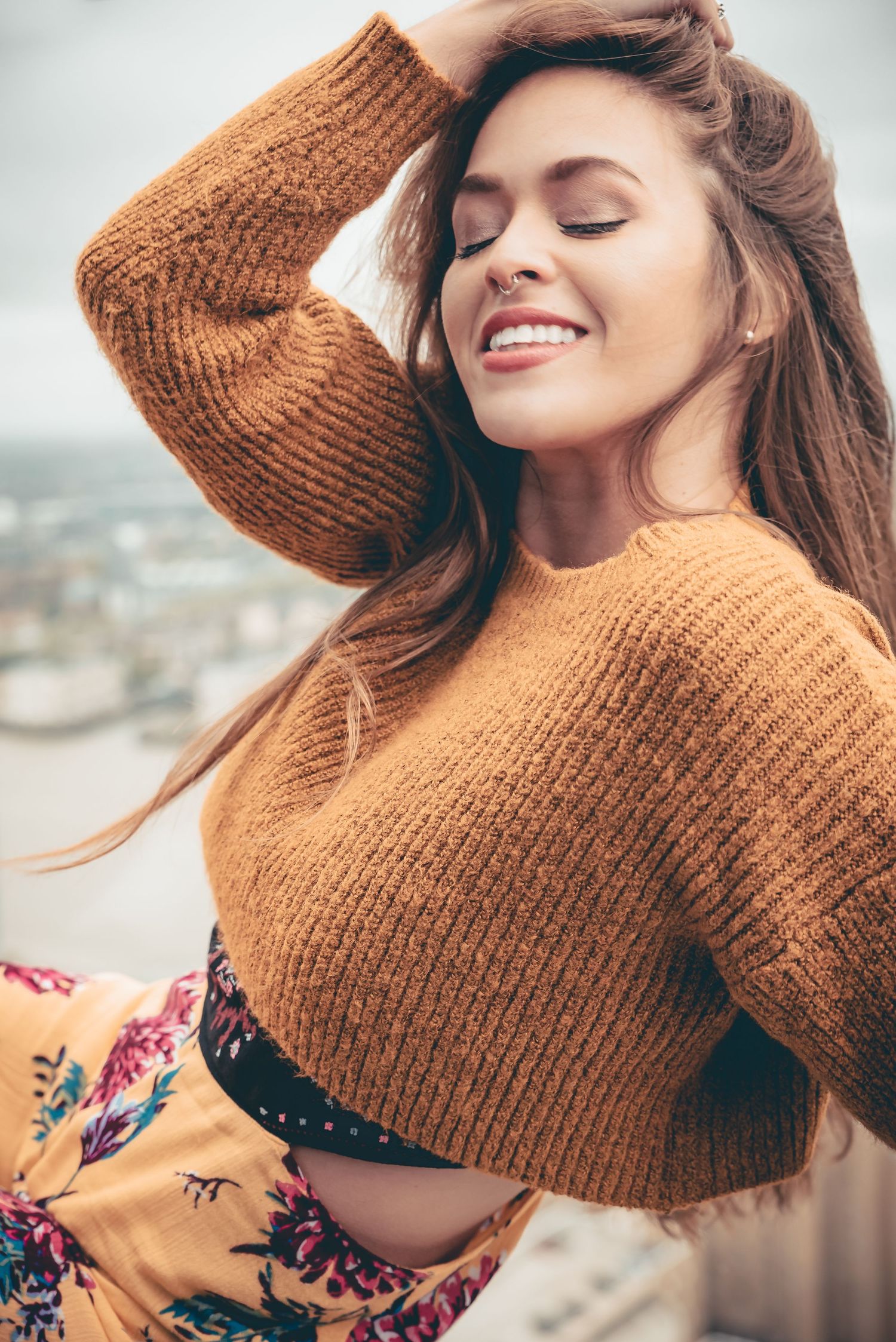 Smiling woman with long hair wearing a brown knit sweater and floral skirt poses outdoors with eyes closed.