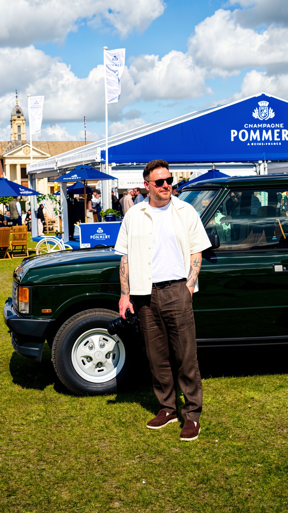 Tattooed man in white shirt and brown trousers stands beside a green vintage car at a Pommery champagne event.