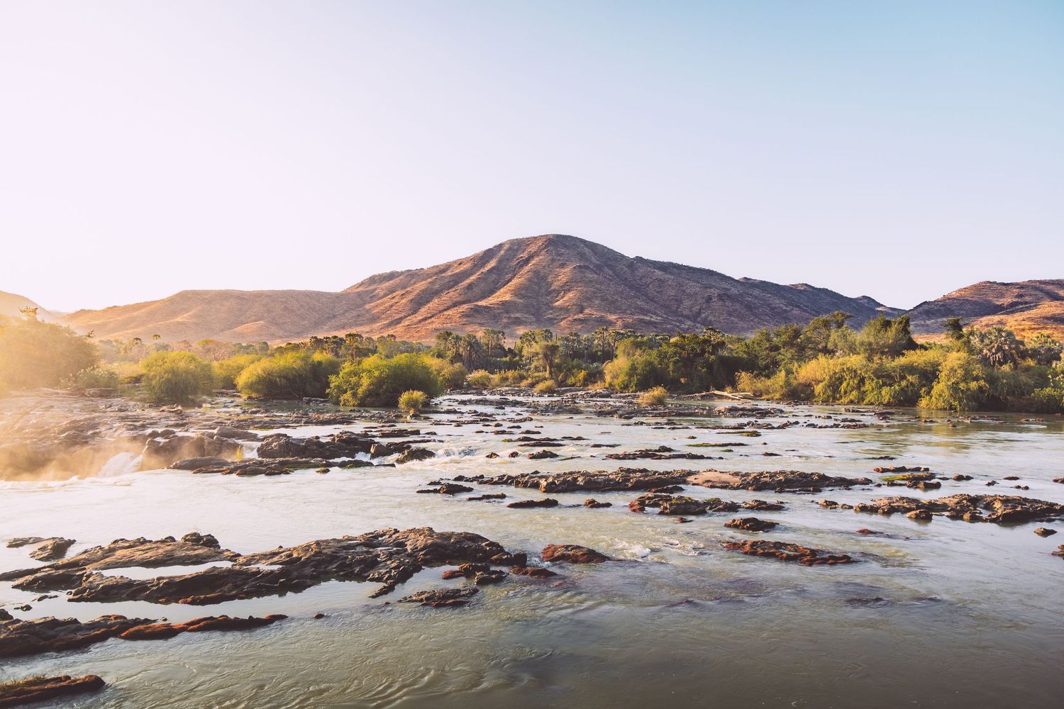 Rocky river flows through arid landscape with lush green trees and rugged brown mountains under a clear blue sky.