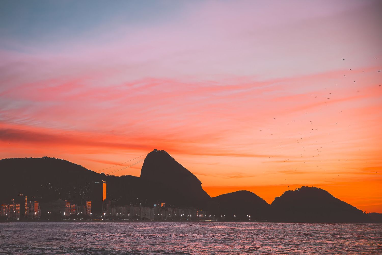 Dramatic silhouette of mountains and Sugar Loaf against a vivid orange and pink sunrise sky in Copacabana, Rio de Janeiro, Brazil.