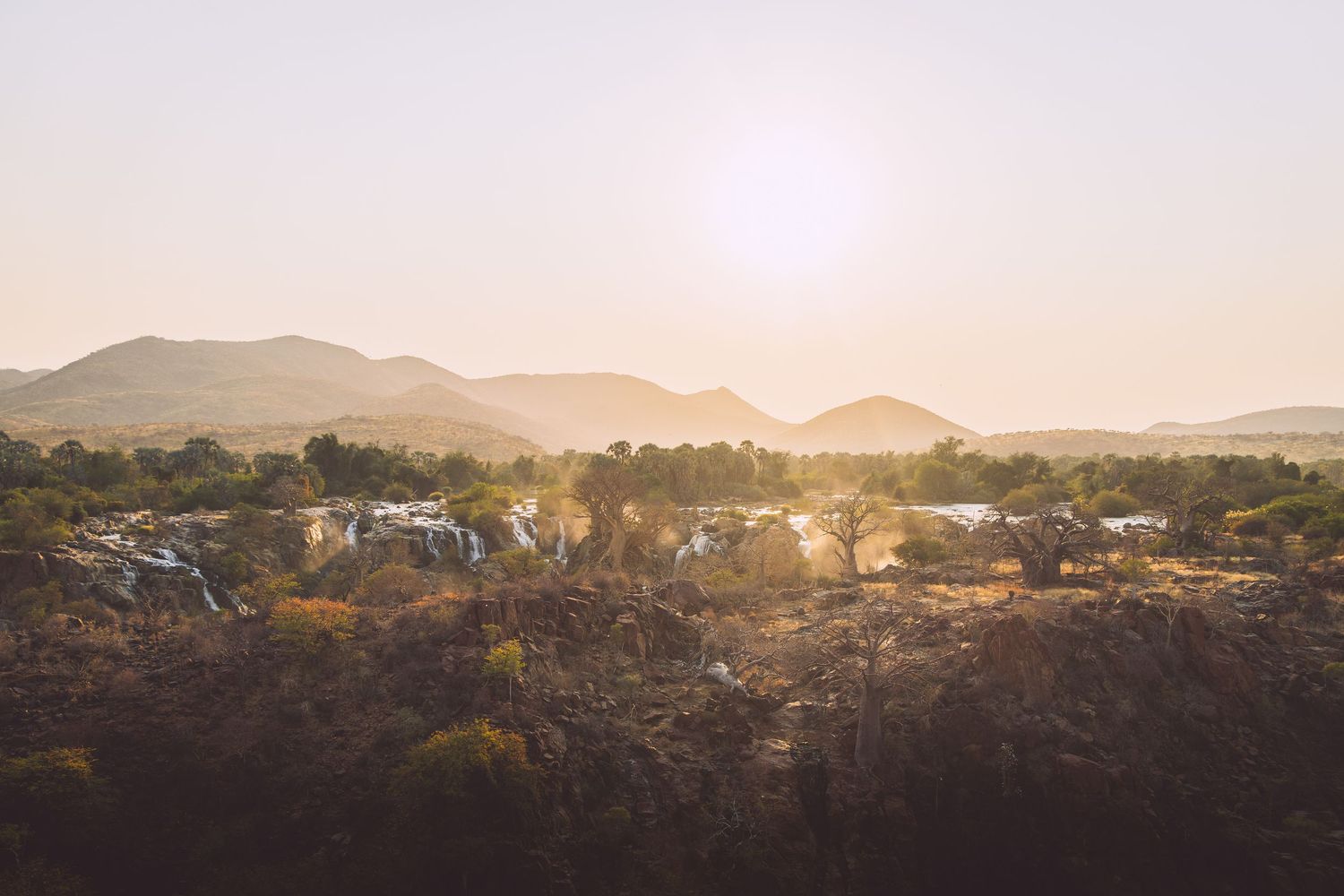 Aerial view of African savanna landscape at golden sunset with mountains, trees, and rocky terrain in warm hazy light.