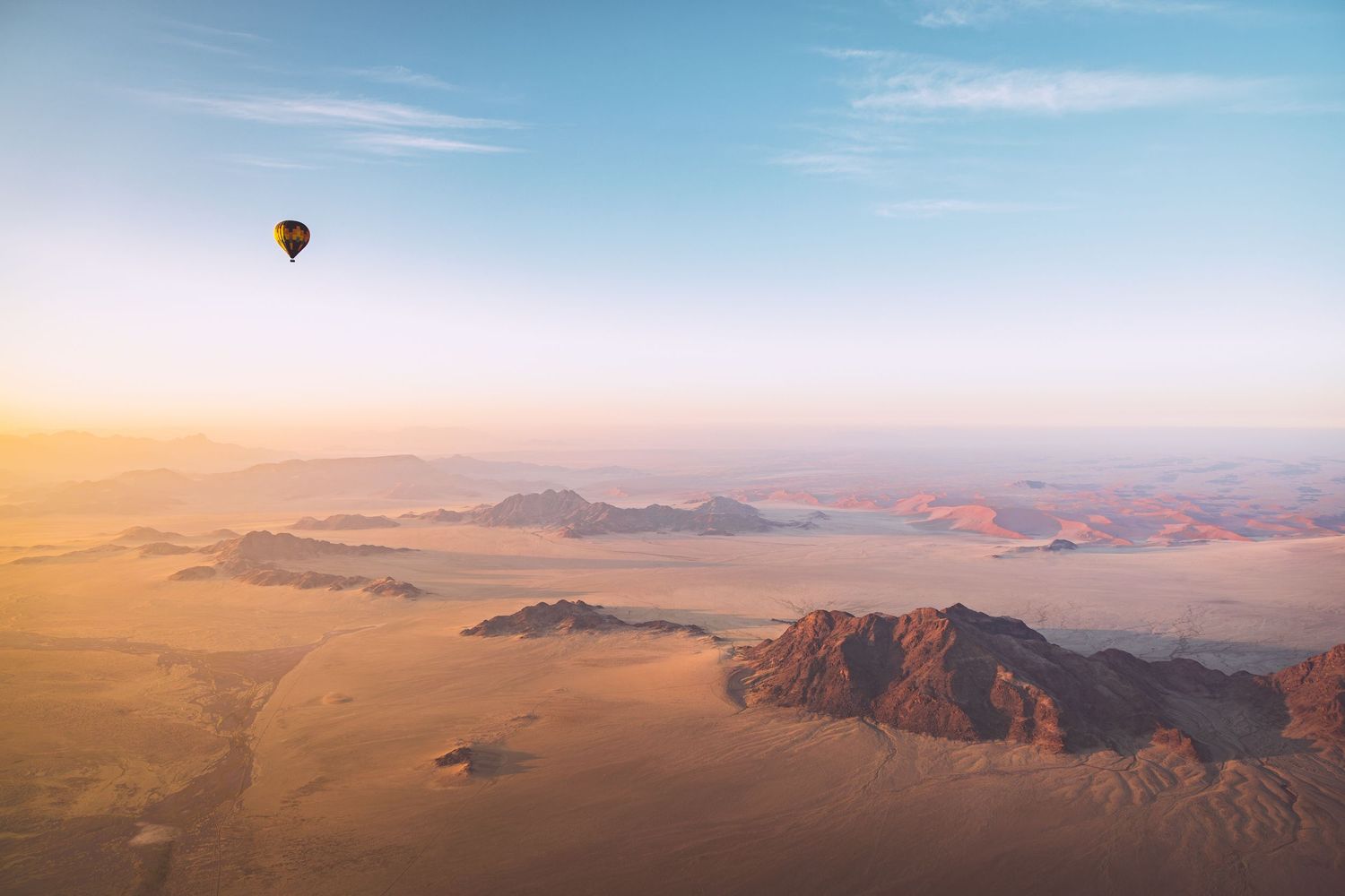 Hot air balloon soaring over vast desert landscape with mountains at golden sunrise.