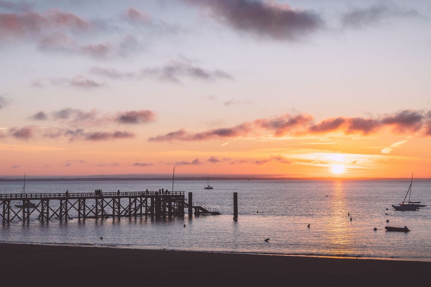 Panoramic view of a coastal sunset with an old wooden pier and boats dotting the horizon in Noirmoutier, France.