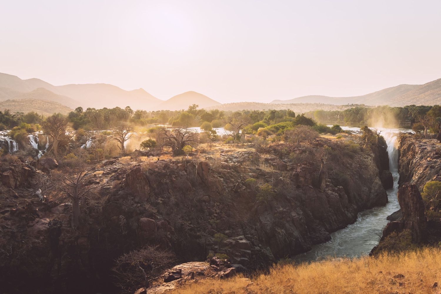 Scenic African canyon with waterfall, rocky cliffs, lush trees, and misty mountains at golden hour.