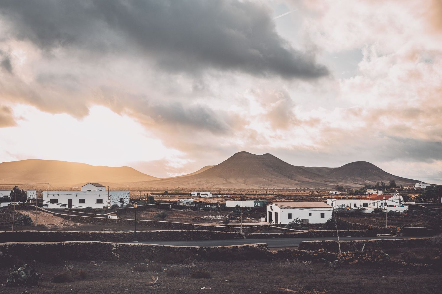 Desert landscape with white buildings and mountains under dramatic cloudy skies in Fuerteventura.