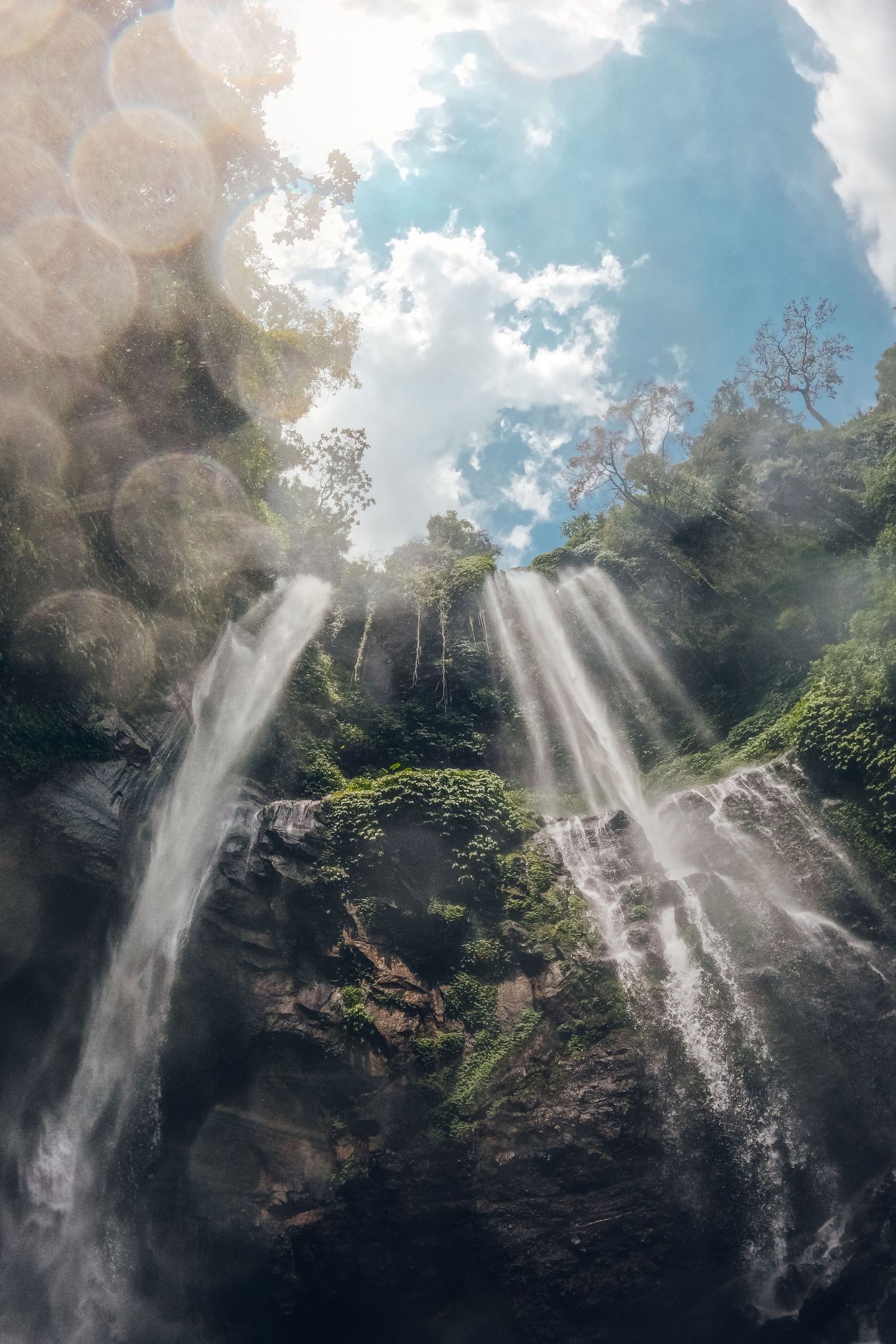 Dramatic waterfall cascading down moss-covered cliffs with sunbeams breaking through the forest canopy in Bali, Indonesia.