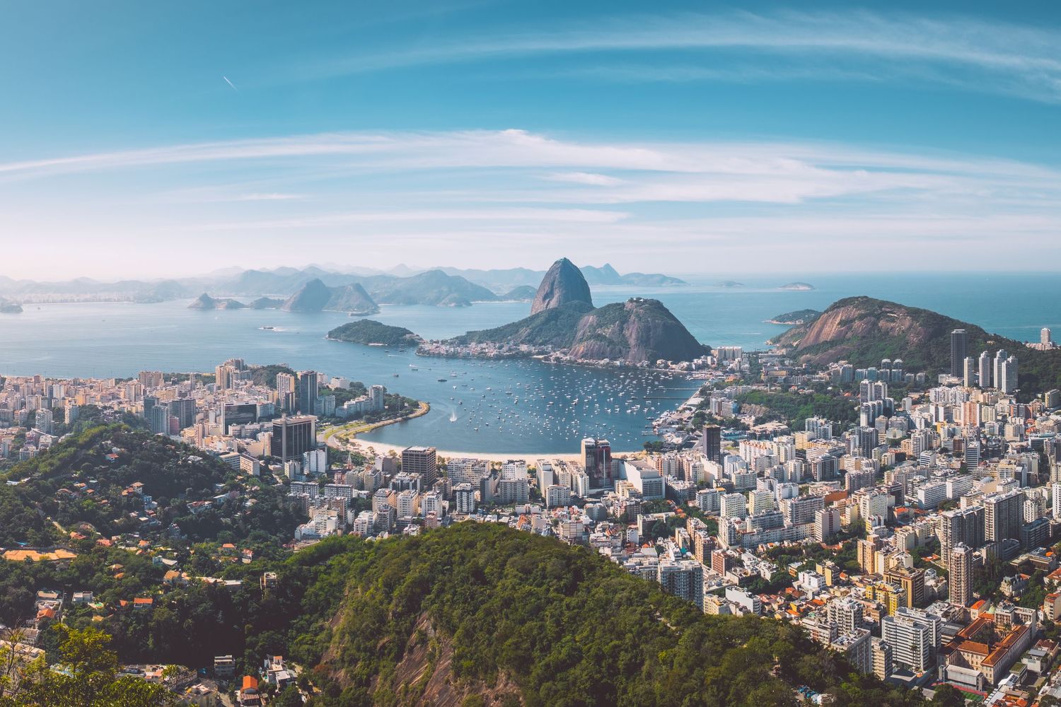 Panoramic cityscape of Rio de Janeiro with mountains and high-rise buildings under blue skies.