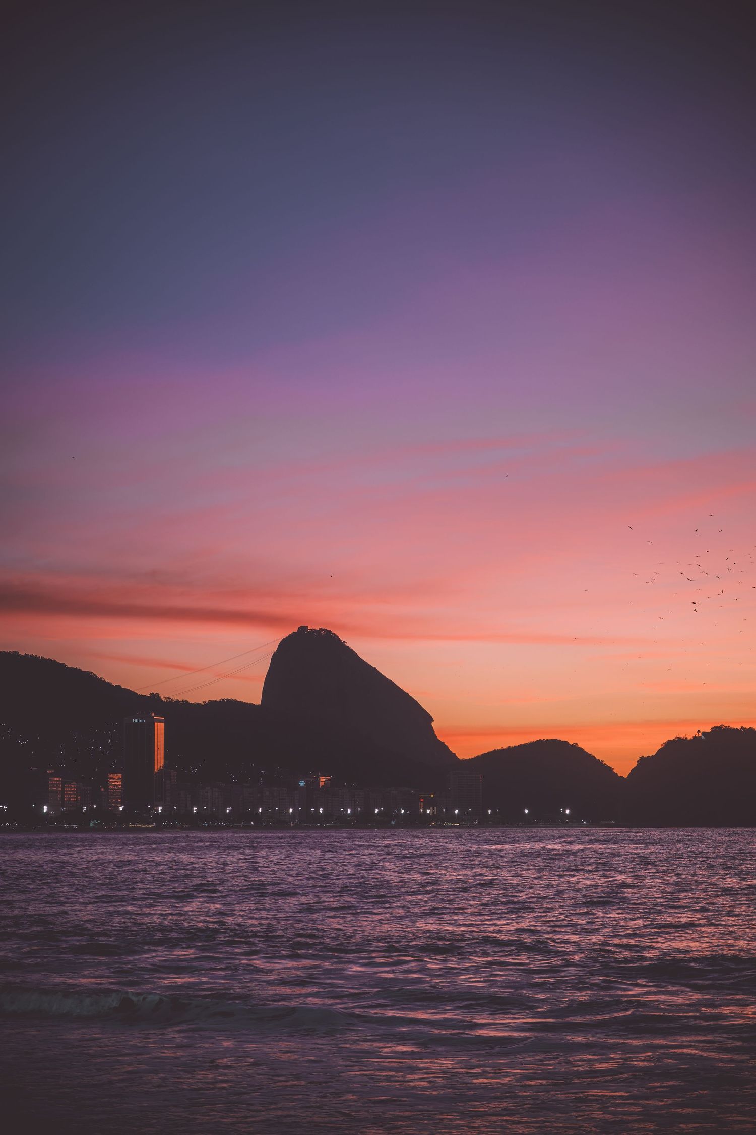 Twilight sunrise seascape with mountain and Sugar Loaf silhouettes and city lights in Copacabana, Rio de Janeiro, Brazil.