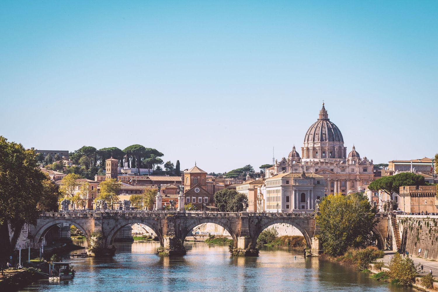St. Peter's Basilica and Sant'Angelo Bridge spanning across Tiber River in Rome under blue sky in Roma, Italy.