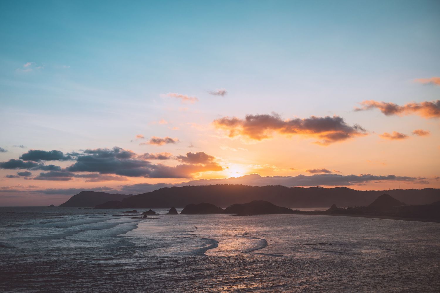 Golden sunset over a rocky coastal landscape with mountains silhouetted against the colorful sky in Lombok, Indonesia.