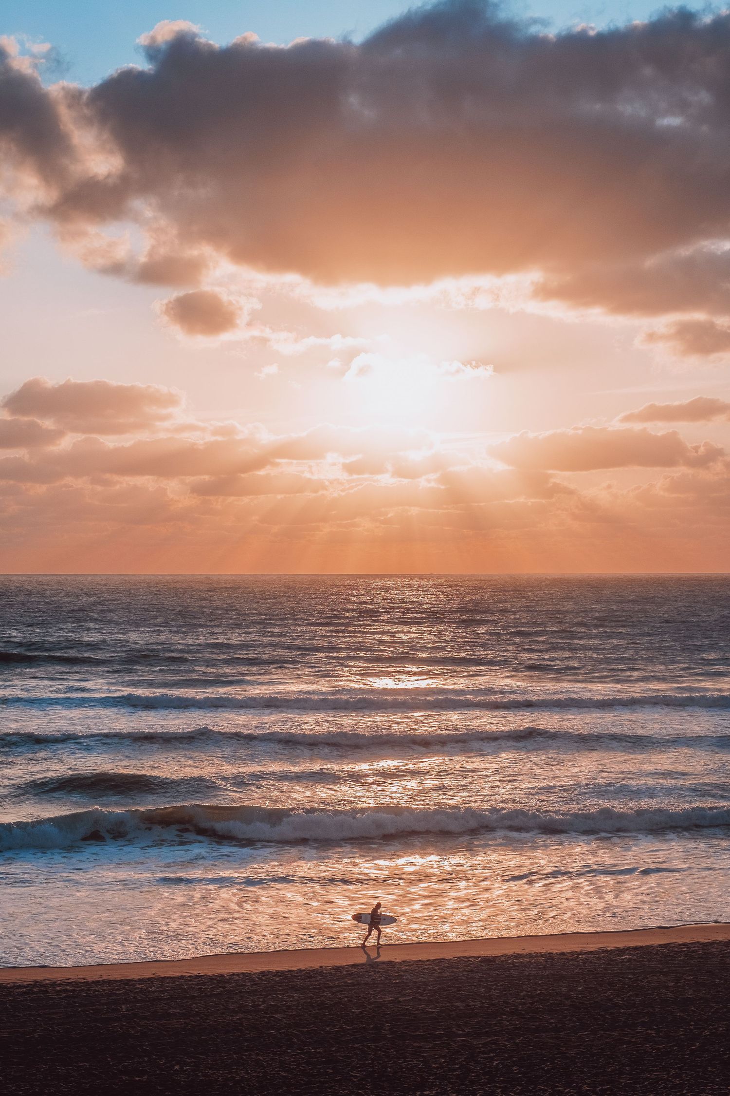 A lone surfer stands on a beach at sunset with dramatic clouds and golden light reflecting on ocean waves in Hourtin, France.