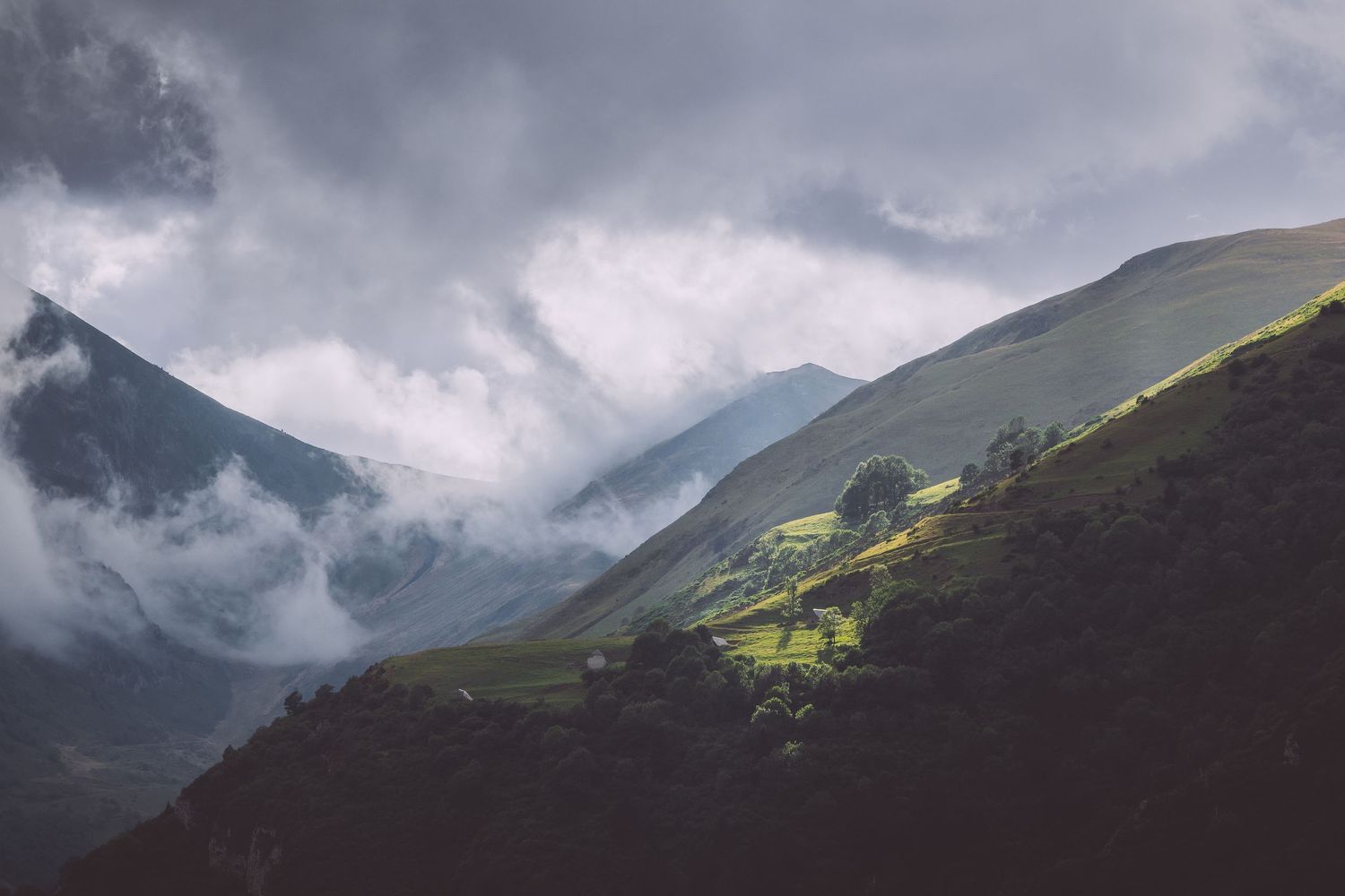 Dramatic storm clouds roll over green mountain peaks in a moody landscape scene in The Pyrénées mountains, France.