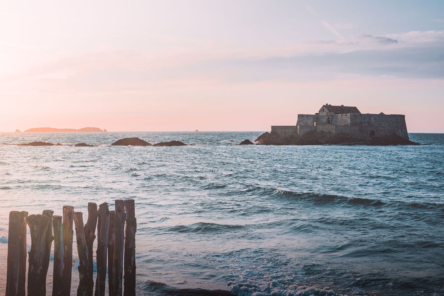 Serene seascape with wooden posts in water and distant fortress silhouette at sunset in Saint-Malo, France.