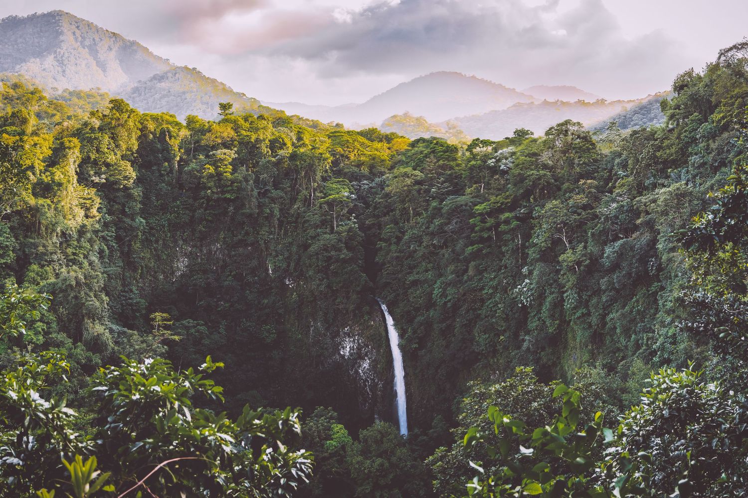 Towering waterfall cascading through lush tropical rainforest valley under cloudy skies in Costa Rica.
