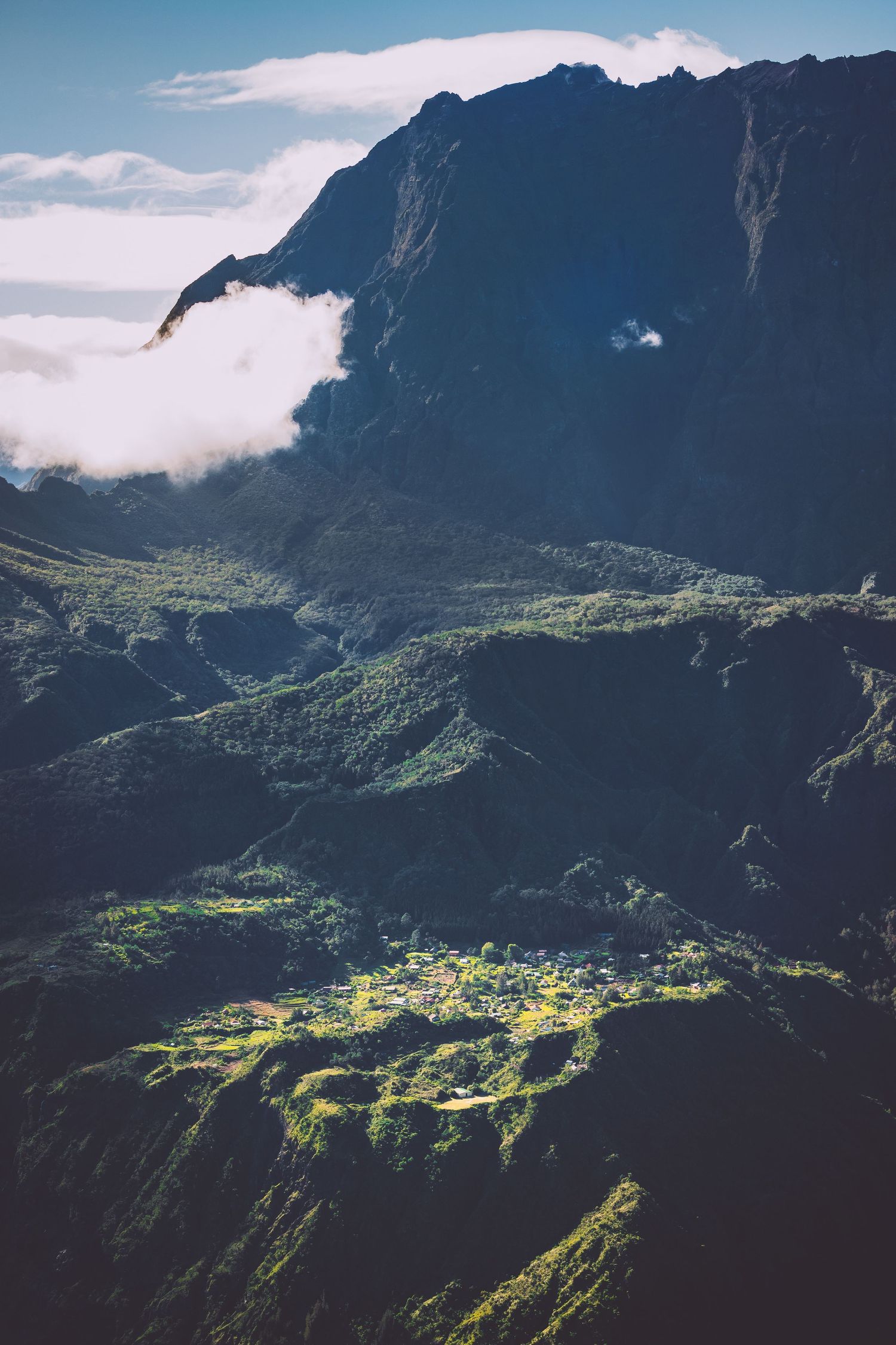 Steep mountain cliffs overlook a small village nestled in a green valley with clouds below in Reunion island.