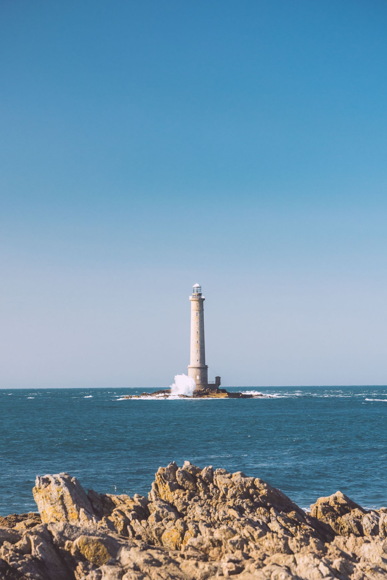 White lighthouse tower standing tall on rocky outcrop in blue ocean waters under clear sky in Cotentin, France.