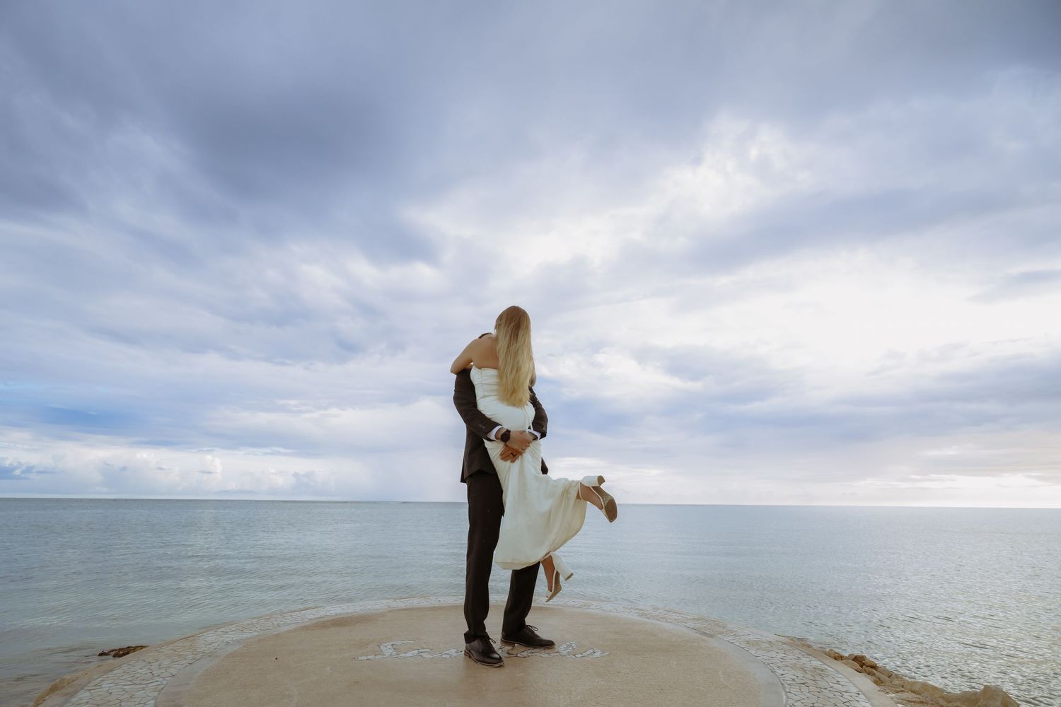 A couple stands on a rocky beach during their wedding photoshoot with dramatic cloudy skies in the background.