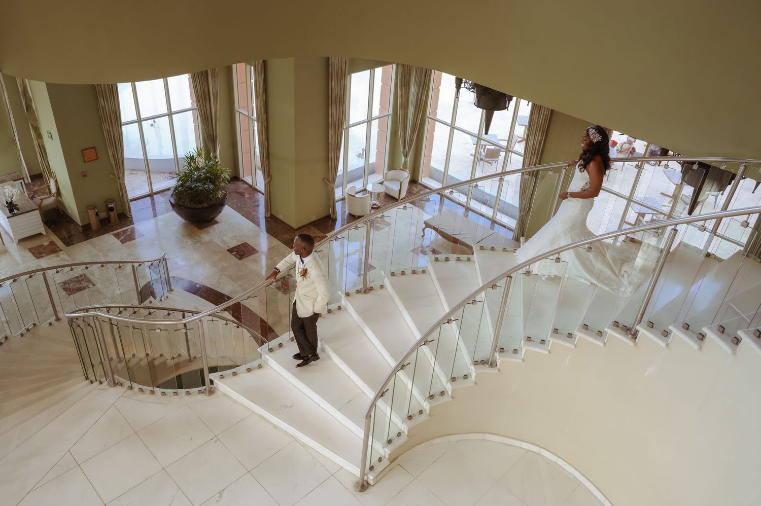 A couple in wedding attire descends a grand staircase with glass railings and natural light.