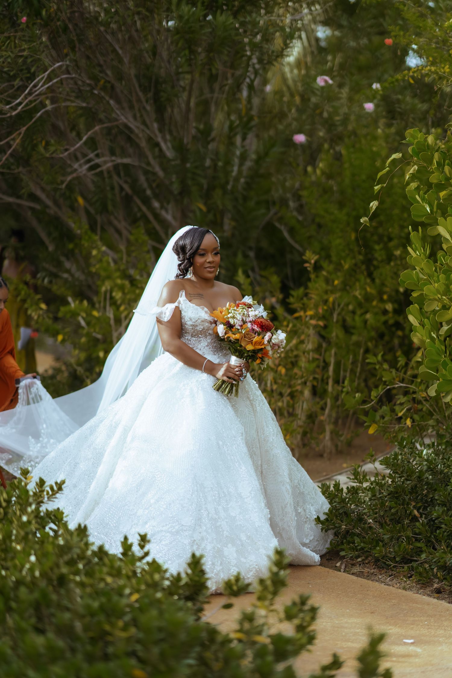 Bride in flowing white wedding dress poses gracefully in a garden setting with colorful flowers and greenery.