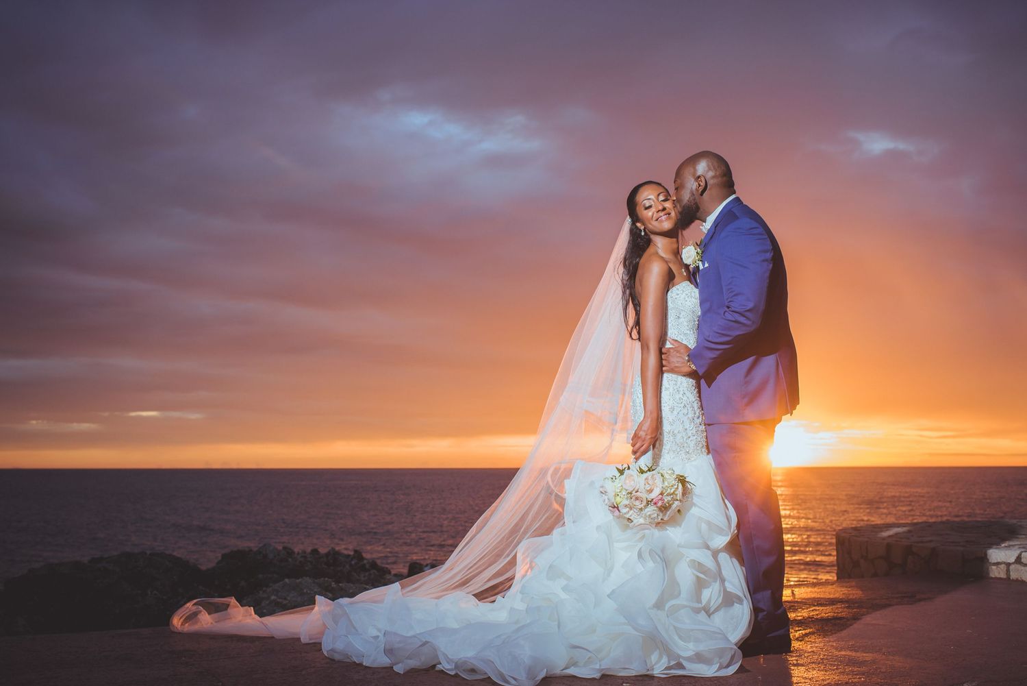 Newlyweds share intimate moment against vibrant sunset sky over ocean.