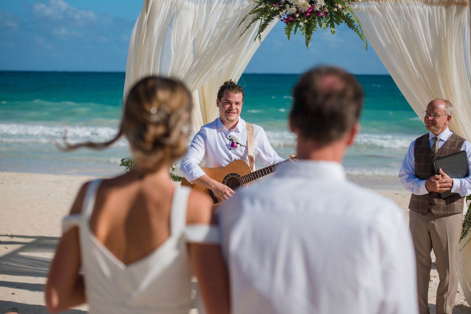 Beach wedding ceremony setup with white fabric arch and chairs against turquoise ocean backdrop.