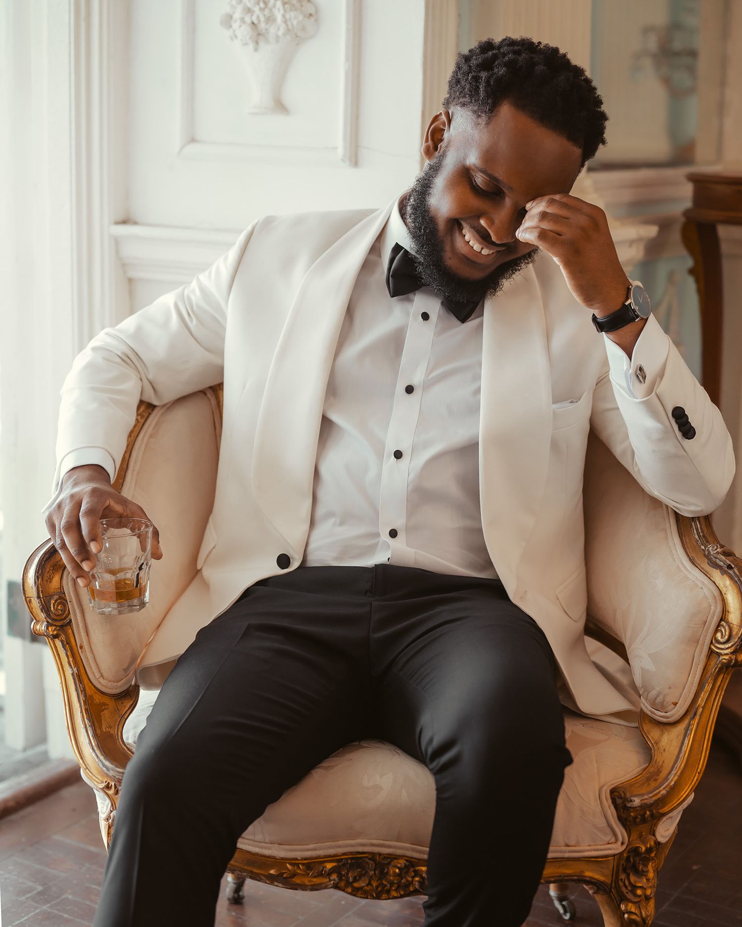 Groom in white tuxedo jacket and black bow tie sits thoughtfully in an ornate gold chair.