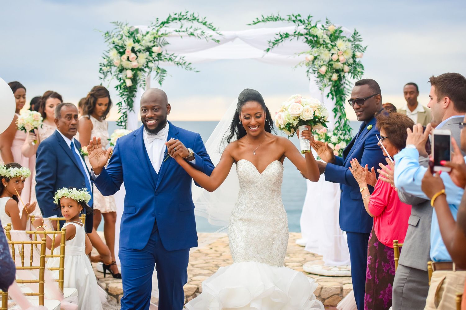 Wedding recessional on stone terrace with guests cheering under floral arch by the sea.