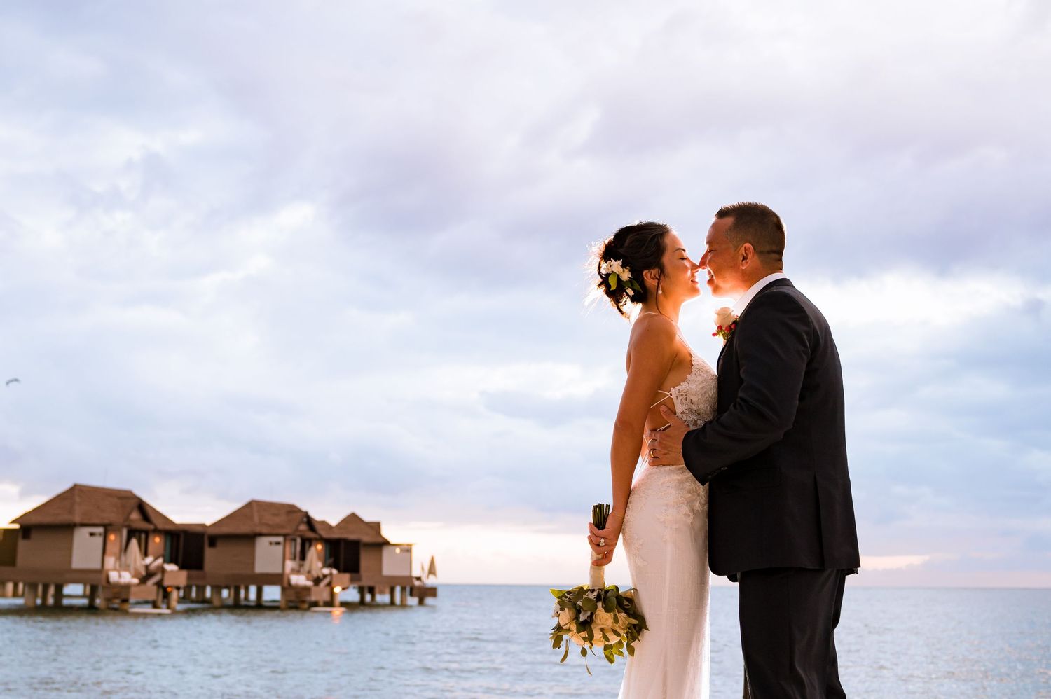 Wedding couple shares intimate moment with overwater bungalows in background at sunset.