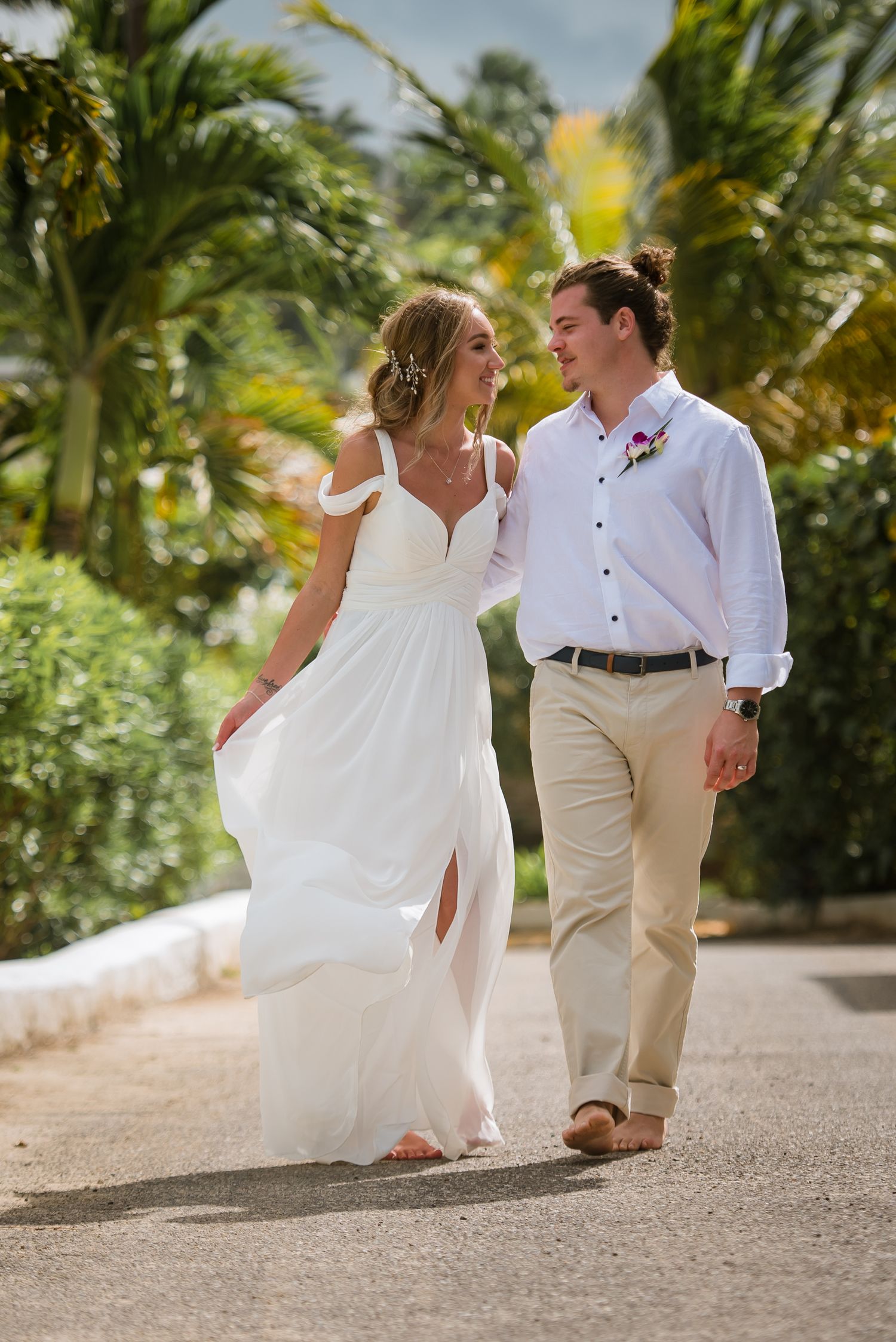 Couple in wedding attire playfully walking and dancing along a palm-lined tropical pathway.